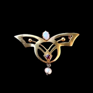 May include: A gold brooch in an Art Nouveau style. It has an oval opal, a small red gemstone, and a dangling pearl. The brooch is symmetrical, with a stylized butterfly or wing design.
