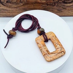 May include: A brown leather cord necklace with a square-shaped pendant made of light brown stone with a smaller square opening in the center. The pendant has a marbled pattern.