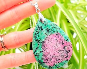 May include: A teardrop-shaped pendant necklace featuring a large, polished gemstone. The stone displays a vibrant mix of green, pink, and black hues, suspended from a delicate silver chain. The pendant is held up by a silver bail.