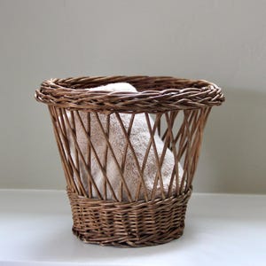 May include: A woven wicker basket with a flared shape, filled with a folded white towel. The basket is a warm brown color and has a decorative, open weave design. The basket is sitting on a white surface.