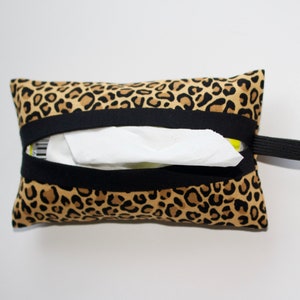 May include: A rectangular tissue holder with a leopard print pattern. The holder is made of fabric with black elastic bands and a black elastic loop. White tissues are visible inside the opening.