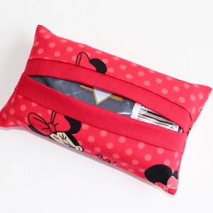 May include: A red tissue holder with a polka dot pattern and a Minnie Mouse graphic. The holder has a red elastic band and is rectangular in shape. The tissue holder is made of fabric.