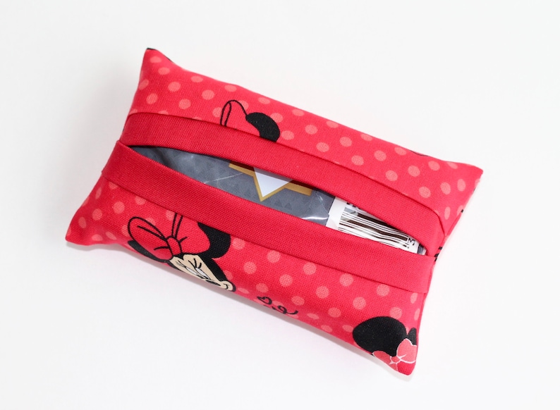 May include: A red tissue holder with a polka dot pattern and a Minnie Mouse graphic. The holder has a red elastic band and is rectangular in shape. The tissue holder is made of fabric.