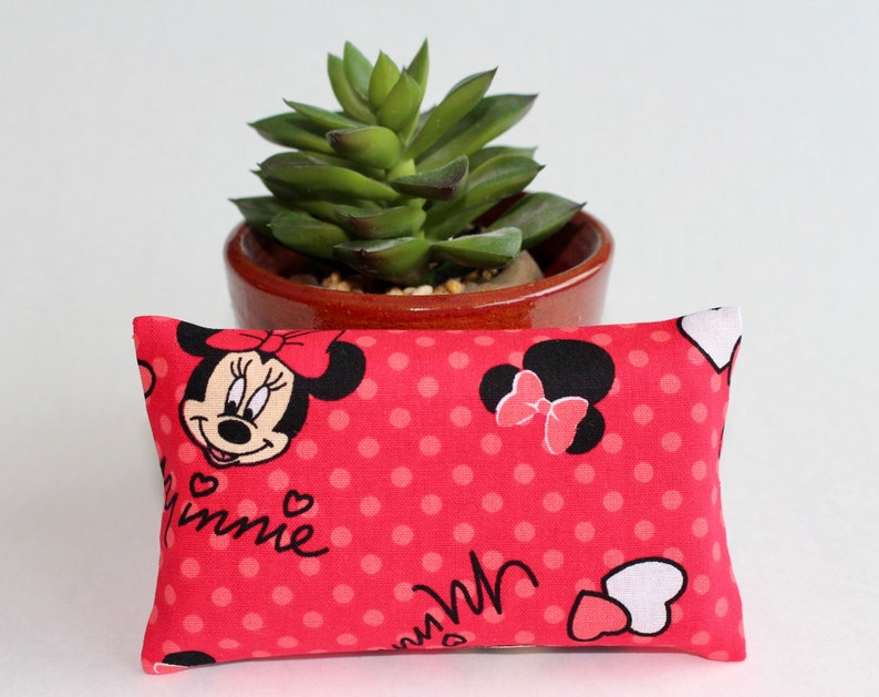May include: A rectangular red fabric item with a Minnie Mouse design. The fabric features a pattern of white polka dots and Minnie Mouse images. The Minnie Mouse images include her face, bow, and name. A small succulent plant in a brown pot is in the background.
