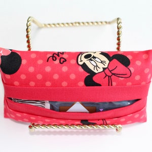 May include: A red fabric tissue holder featuring a Minnie Mouse design with a red bow and polka dots. The holder is displayed on a gold-colored metal stand. The tissue holder has a slot for easy access to tissues.