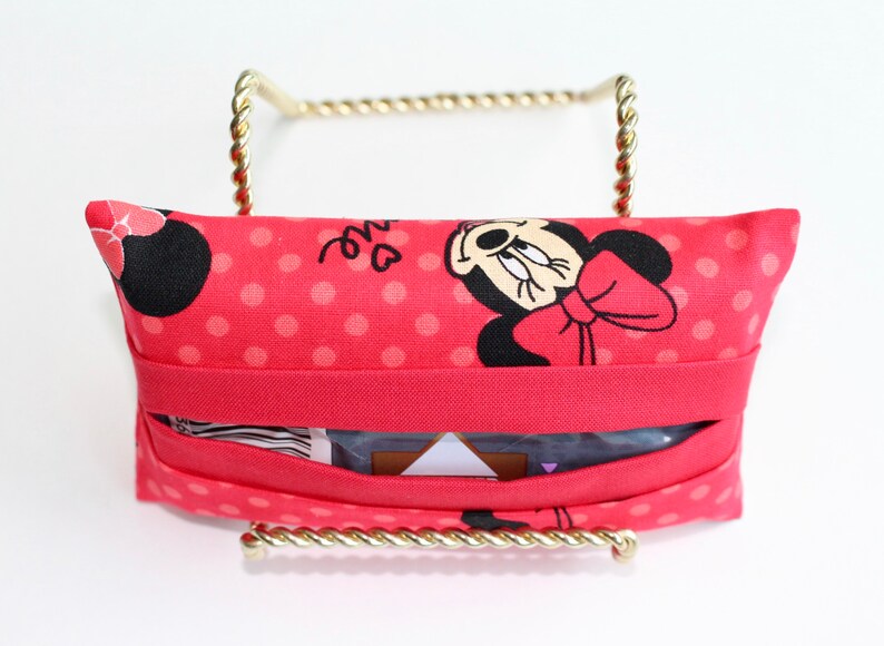 May include: A red fabric tissue holder featuring a Minnie Mouse design with a red bow and polka dots. The holder is displayed on a gold-colored metal stand. The tissue holder has a slot for easy access to tissues.