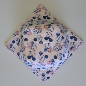 Może przedstawiać: Kwadratowa, pikowana osłona na miskę z powtarzającym się wzorem Myszki Miki i Myszki Minnie. Wzór zawiera niebieskie i różowe akcenty na jasnoróżowym tle. Osłona ma pomieścić miskę i chronić dłonie przed gorącem.