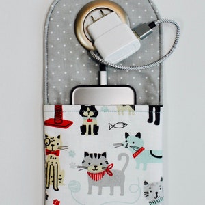 May include: A gray fabric charging station with a white polka dot pattern and a pocket featuring a cat print. A white charger and cable are connected to a device inside the pocket.