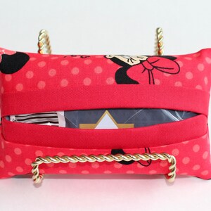 May include: A red tissue box cover with a Minnie Mouse pattern, featuring black silhouettes and white polka dots. The cover is displayed on a gold-colored metal stand with a twisted rope design.
