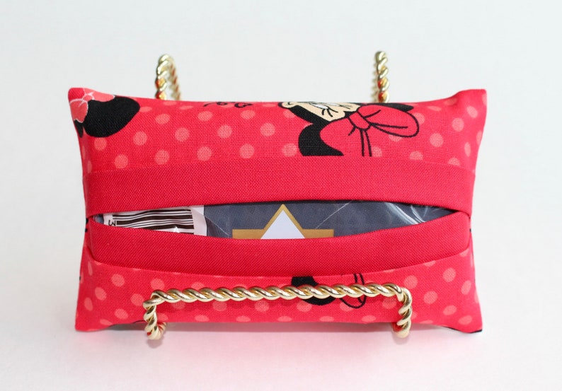 May include: A red tissue box cover with a Minnie Mouse pattern, featuring black silhouettes and white polka dots. The cover is displayed on a gold-colored metal stand with a twisted rope design.