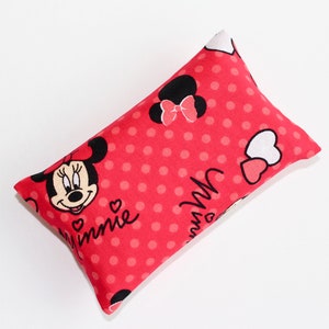 May include: A rectangular, red pillow featuring a pattern of white polka dots and Minnie Mouse graphics. The pillow has a Minnie Mouse face and the word "Minnie" printed on it. The pillow is filled and has a soft appearance.