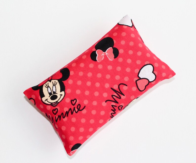May include: A rectangular, red pillow featuring a pattern of white polka dots and Minnie Mouse graphics. The pillow has a Minnie Mouse face and the word "Minnie" printed on it. The pillow is filled and has a soft appearance.