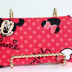 May include: A red rectangular pillow with a Minnie Mouse design, featuring her face, polka dots, and the name "Minnie" in cursive. The pillow is displayed on a gold-colored metal stand.
