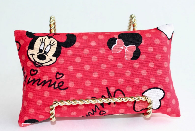 May include: A red rectangular pillow with a Minnie Mouse design, featuring her face, polka dots, and the name "Minnie" in cursive. The pillow is displayed on a gold-colored metal stand.