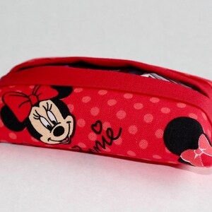 May include: Red fabric pouch with a Minnie Mouse design. The pouch features a red polka dot pattern and a Minnie Mouse graphic on the front. The pouch has a red zipper and is rectangular in shape.