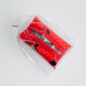 May include: A small, white, sheer drawstring bag containing several red fabric items. The red fabric has a polka dot pattern and a Minnie Mouse graphic. The bag is tied with a white ribbon.