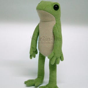May include: A plush frog toy with a light green exterior and a beige belly. The frog has large black eyes and stands on two legs with four-toed feet. The text "car2in-bitz Arts & Crafts 2025" is visible on the chest of the plush frog figure.
