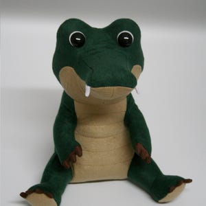 Puede incluir: Un peluche de cocodrilo verde y beige. El cocodrilo tiene ojos grandes y redondos, dientes blancos y garras marrones. El juguete está sentado erguido, con las piernas extendidas. Hecho de tela suave.