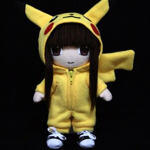 May include: A plush toy doll wearing a yellow Pikachu costume with black hair and a zipper on the front. The doll is wearing black and white sneakers.
