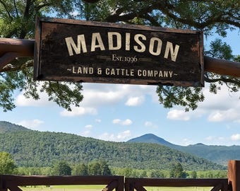 May include: A weathered wooden sign with the name "MADISON" in large white letters, "Est. 1996" below, and "LAND & CATTLE COMPANY" at the bottom. The sign hangs above a wooden gate with a mountain range in the background.