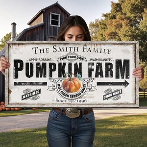 May include: A rustic wooden sign with the text "The Smith Family Pumpkin Farm" in black lettering. The sign includes illustrations of a pumpkin, arrows, and the words "Hayrides", "Hot Cider Served Daily", and "Corn Maze". The sign is held by a person in a rural setting.