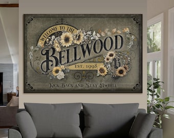May include: A rectangular sign with the text "WELCOME TO THE BELLWOOD EST. 1998" in a vintage style. The sign features sunflowers and floral accents in shades of yellow, cream, and green. The phrase "KICK BACK AND STAY AWHILE" is at the bottom.