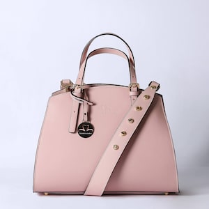 Pu&ograve; includere: Una borsa a mano in pelle rosa chiaro con una fibbia dorata e una tracolla rimovibile. La borsa ha un logo a forma di cuore con il testo "Victoria London" sul davanti.