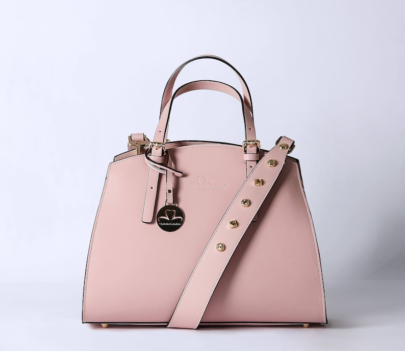 Pu&ograve; includere: Una borsa a mano in pelle rosa chiaro con una fibbia dorata e una tracolla rimovibile. La borsa ha un logo a forma di cuore con il testo "Victoria London" sul davanti.