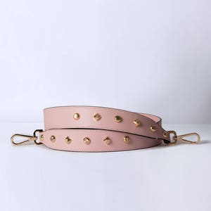 Pu&ograve; includere: Una tracolla in pelle rosa con borchie dorate. La tracolla ha due fibbie metalliche.