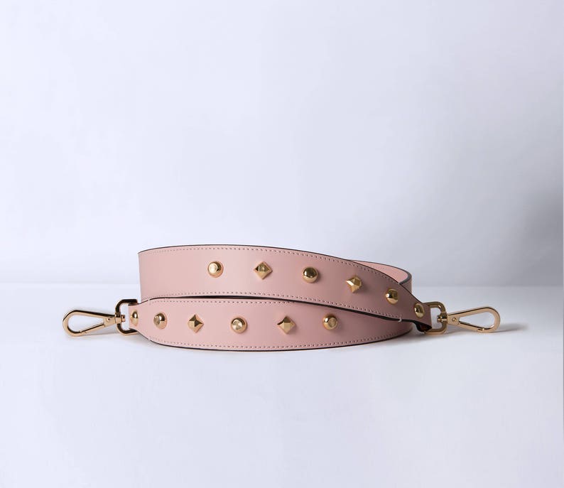 Pu&ograve; includere: Una tracolla in pelle rosa con borchie dorate. La tracolla ha due fibbie metalliche.