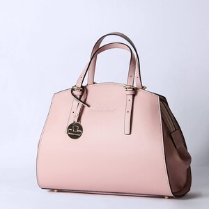 Pu&ograve; includere: Una borsa a mano in pelle rosa chiaro con una fibbia dorata e un piccolo ciondolo dorato. La borsa ha un manico superiore e una tracolla rimovibile.