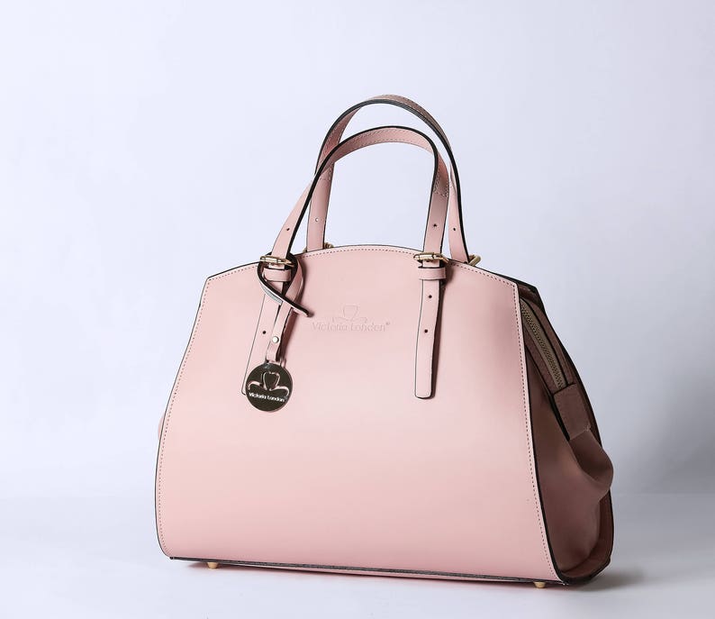Pu&ograve; includere: Una borsa a mano in pelle rosa chiaro con una fibbia dorata e un piccolo ciondolo dorato. La borsa ha un manico superiore e una tracolla rimovibile.