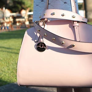 Pu&ograve; includere: Una borsa a mano in pelle rosa chiaro con una fibbia dorata e una tracolla con borchie. La borsa ha un logo rotondo in metallo con le parole "Pink & Love" su di esso.