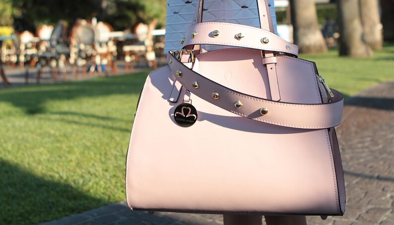 Pu&ograve; includere: Una borsa a mano in pelle rosa chiaro con una fibbia dorata e una tracolla con borchie. La borsa ha un logo rotondo in metallo con le parole "Pink & Love" su di esso.