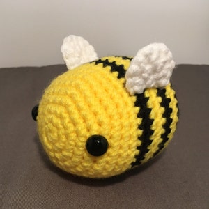 May include: A crocheted bee toy with a round body. The bee is yellow with black stripes and white wings. It has two black bead eyes. The toy is handmade and appears soft.