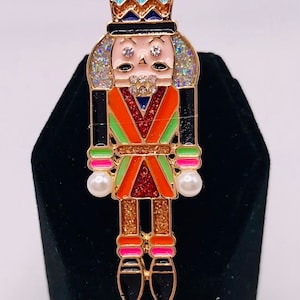 May include: A gold-tone nutcracker brooch with a red, green, and orange patterned design. The nutcracker has a black hat with a blue and orange chevron pattern, a white face with rhinestones, and pearl accents.