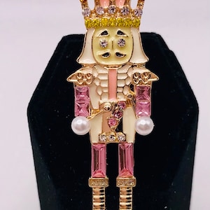 May include: A decorative brooch in the shape of a nutcracker soldier. The brooch features a pink and white uniform, a gold crown with yellow and clear rhinestones, and pink and clear rectangular gems. Two faux pearls are on the sides.