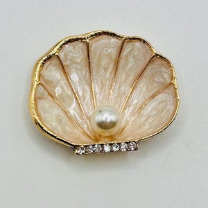 May include: A seashell-shaped brooch with a gold-tone border. The interior features a pearlescent, fan-like design with a central pearl. A row of small, clear crystals adorns the bottom edge, adding sparkle to this jewelry piece.