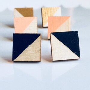 May include: A set of six geometric wooden earrings. Each earring is a square with a diagonal line dividing it into two triangles. The triangles are painted in different colors, including black, white, pink, and gold.