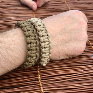 May include: Two brown paracord bracelets, one slightly wider than the other, are worn on a person's wrist. The bracelets are made with a woven pattern.
