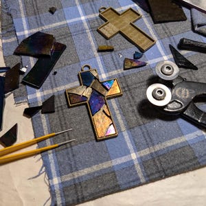 Peut inclure: Gros plan sur une croix en vitrail avec un cadre dor&eacute;. La croix est compos&eacute;e de petits morceaux de verre dans des tons de bleu, violet et or. La croix est entour&eacute;e d'outils pour travailler le vitrail, notamment une paire de coupe-verre et une pince.