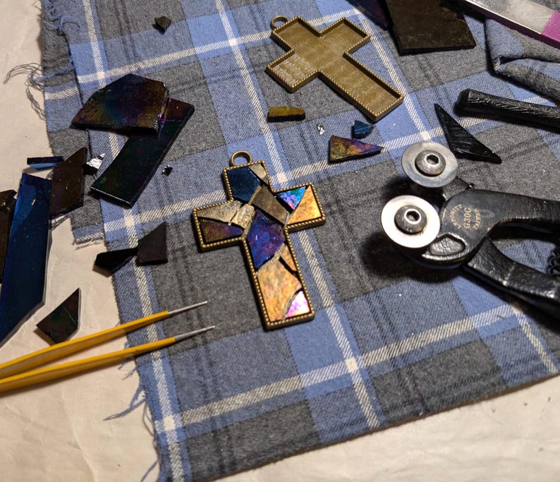 Peut inclure: Gros plan sur une croix en vitrail avec un cadre dor&eacute;. La croix est compos&eacute;e de petits morceaux de verre dans des tons de bleu, violet et or. La croix est entour&eacute;e d'outils pour travailler le vitrail, notamment une paire de coupe-verre et une pince.