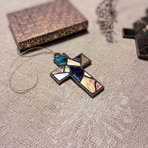 Peut inclure: Un pendentif en forme de croix en verre color&eacute; avec un cadre dor&eacute;. La croix est compos&eacute;e de petits morceaux de verre dans des tons de bleu, violet et marron. Le pendentif est suspendu &agrave; une fine cha&icirc;ne dor&eacute;e.