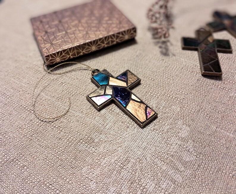 Peut inclure: Un pendentif en forme de croix en verre color&eacute; avec un cadre dor&eacute;. La croix est compos&eacute;e de petits morceaux de verre dans des tons de bleu, violet et marron. Le pendentif est suspendu &agrave; une fine cha&icirc;ne dor&eacute;e.