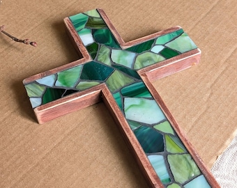 May include: A cross-shaped decorative object featuring a mosaic of various green and white glass pieces set within a wooden frame with a reddish-brown finish. The glass fragments vary in shade from deep forest green to pale mint and white.
