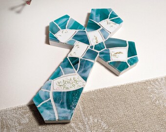 Peut inclure: Une croix décorative présentant un motif en mosaïque composé d'éclats de verre bleu sarcelle et de pièces en céramique blanche. Certains fragments blancs présentent de délicats motifs floraux dans des tons verts et bruns sur une base blanche.