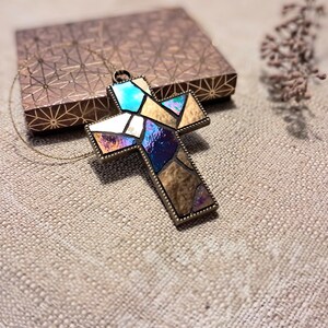 Peut inclure: Un collier dor&eacute; avec un pendentif en forme de croix en verre color&eacute;. La croix est faite de morceaux de verre iridescent dans les tons bleus, violets et dor&eacute;s.