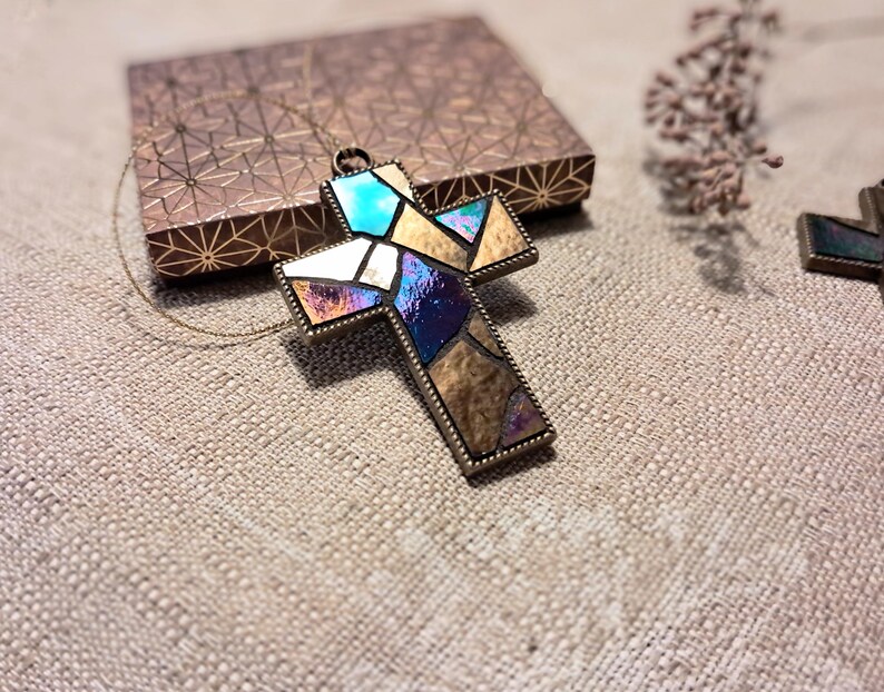 Peut inclure: Un collier dor&eacute; avec un pendentif en forme de croix en verre color&eacute;. La croix est faite de morceaux de verre iridescent dans les tons bleus, violets et dor&eacute;s.