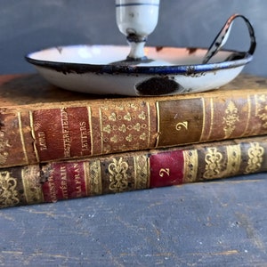 Op de afbeelding: Twee antieke boeken die op elkaar gestapeld zijn. Het bovenste boek is getiteld "Lord Chesterfield's Letters" en het onderste boek is getiteld "Augustus the Fair, La Fran". Een witte emaille kaarsenstandaard met een blauwe rand staat op de boeken.