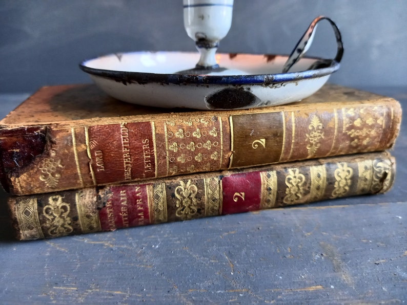 Op de afbeelding: Twee antieke boeken die op elkaar gestapeld zijn. Het bovenste boek is getiteld "Lord Chesterfield's Letters" en het onderste boek is getiteld "Augustus the Fair, La Fran". Een witte emaille kaarsenstandaard met een blauwe rand staat op de boeken.